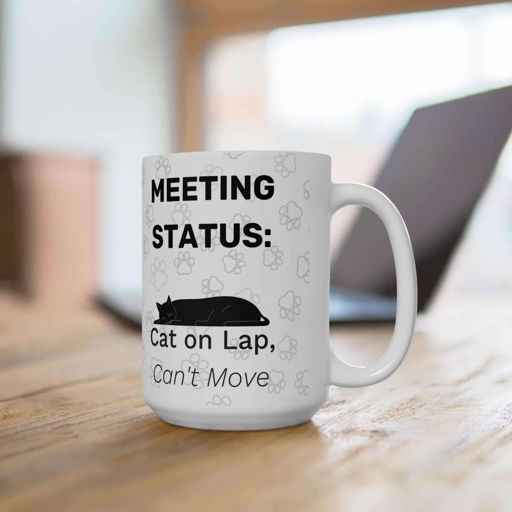 White ceramic mug displaying "Meeting Status: Cat on Lap, Can't Move" text with black silhouette of sleeping cat and paw print pattern, positioned on wooden desk with laptop in background, part of Cat on Lap Meeting Status collection.