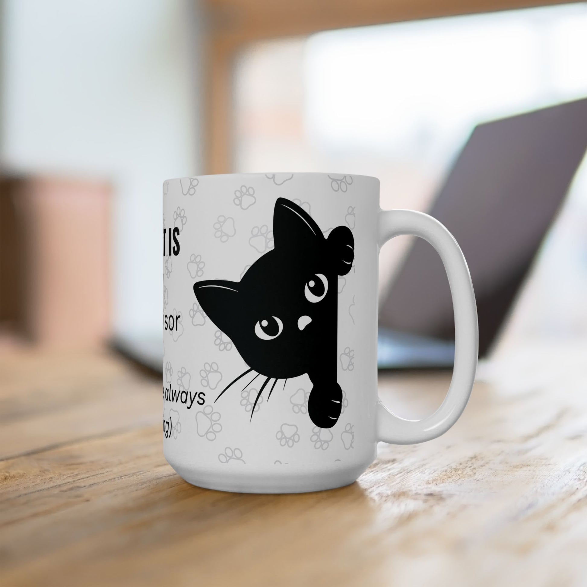 White ceramic mug featuring "The Cat Is My Supervisor" design with a playful black cat silhouette peeking from the side, set against a paw print pattern background, photographed on wooden desk with laptop in background, ideal for remote workers.