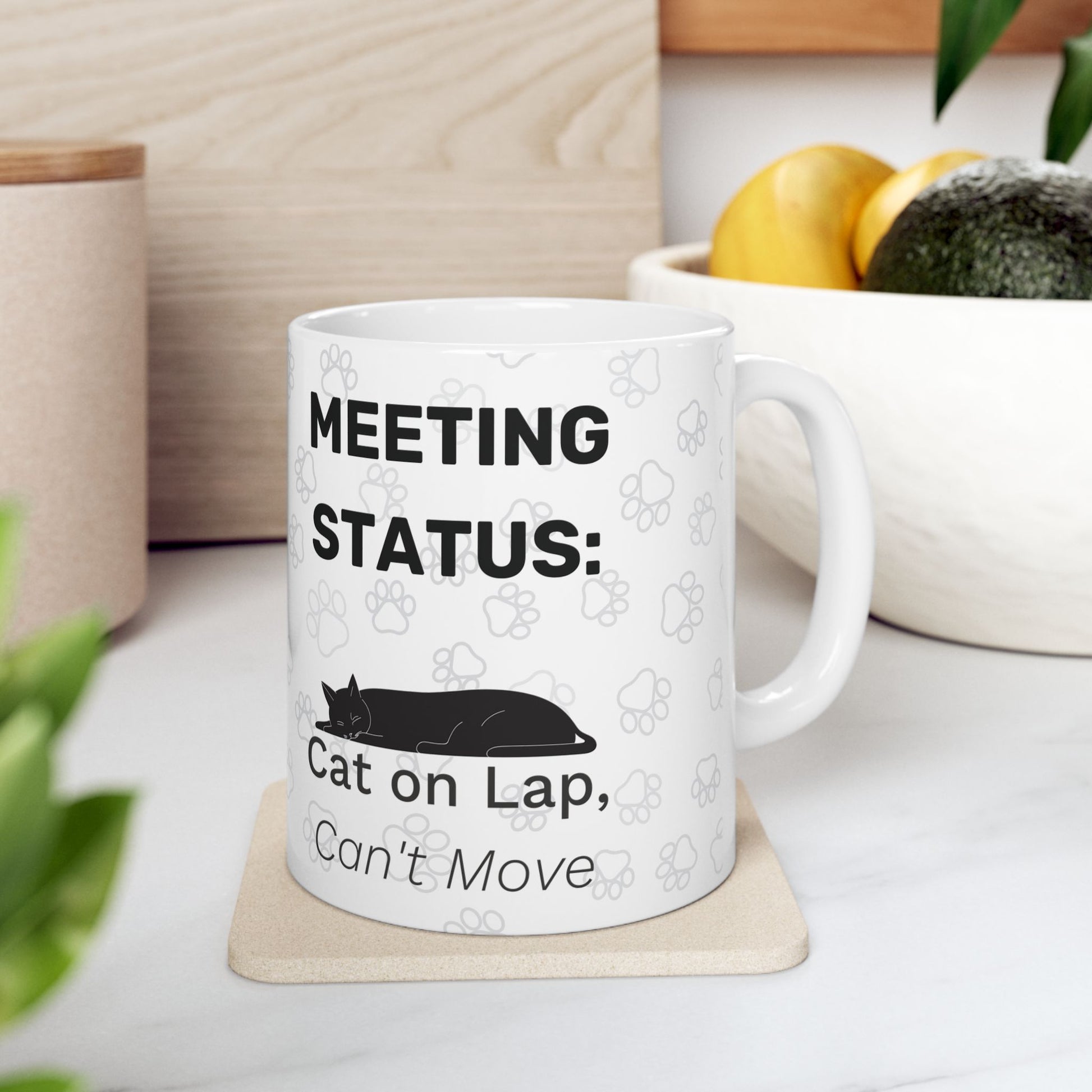 White ceramic mug displaying "Meeting Status: Cat on Lap, Can't Move" text with black cat silhouette and paw print pattern, shown on beige coaster with fruit bowl in background. Perfect for remote workers with feline supervisors.