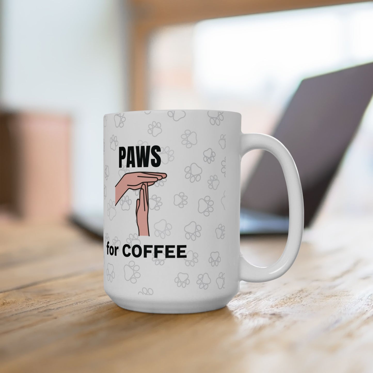 White ceramic mug displaying "Paws for Coffee" design featuring a hand gesture mimicking cat paws against a background of subtle paw print patterns, shown on wooden surface with laptop in background. The Paws for Coffee Cat Lover Mug combines playful design with practical function.