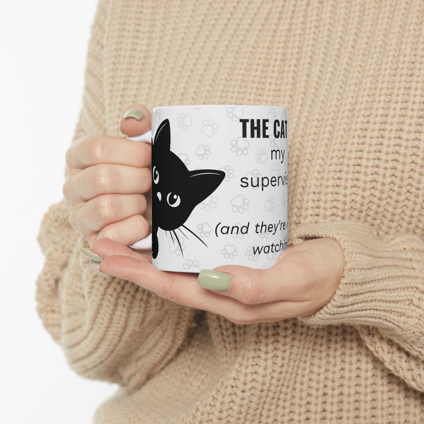 THE CAT IS MY SUPERVISOR work from home mug featuring a white ceramic design with black cat silhouette and paw prints. Hands holding mug wearing beige sweater showing dual-sided design with humorous text about feline supervision, available in 11oz and 15oz sizes.