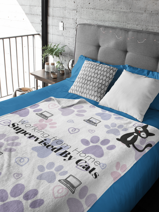 Working from Home Supervised by Cats Sherpa Fleece Blanket displayed on blue bedding, featuring playful paw print pattern in purple and pink, laptop icons, and cat illustrations on white background. Cozy throw blanket perfect for remote workers and pet lovers.