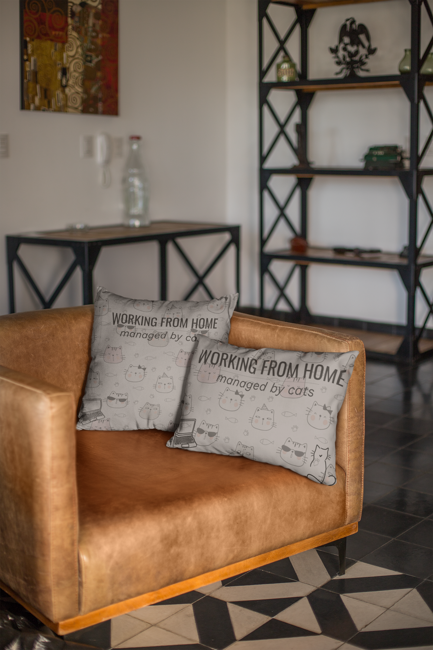 Working From Home: Managed By Cats decorative throw pillows displayed on tan leather couch, featuring cute cat doodles and text design in gray color, shown in modern home office setting with industrial-style shelving in background.