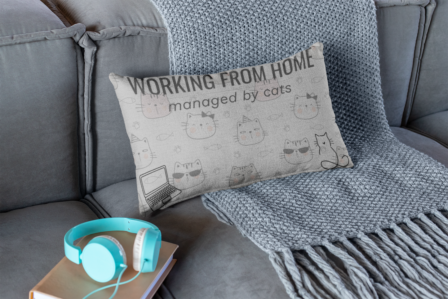 Working From Home: Managed By Cats decorative throw pillow in light gray featuring cute cat face doodles, displayed on gray couch with turquoise headphones and knit blanket, perfect for home office decor and remote workers.