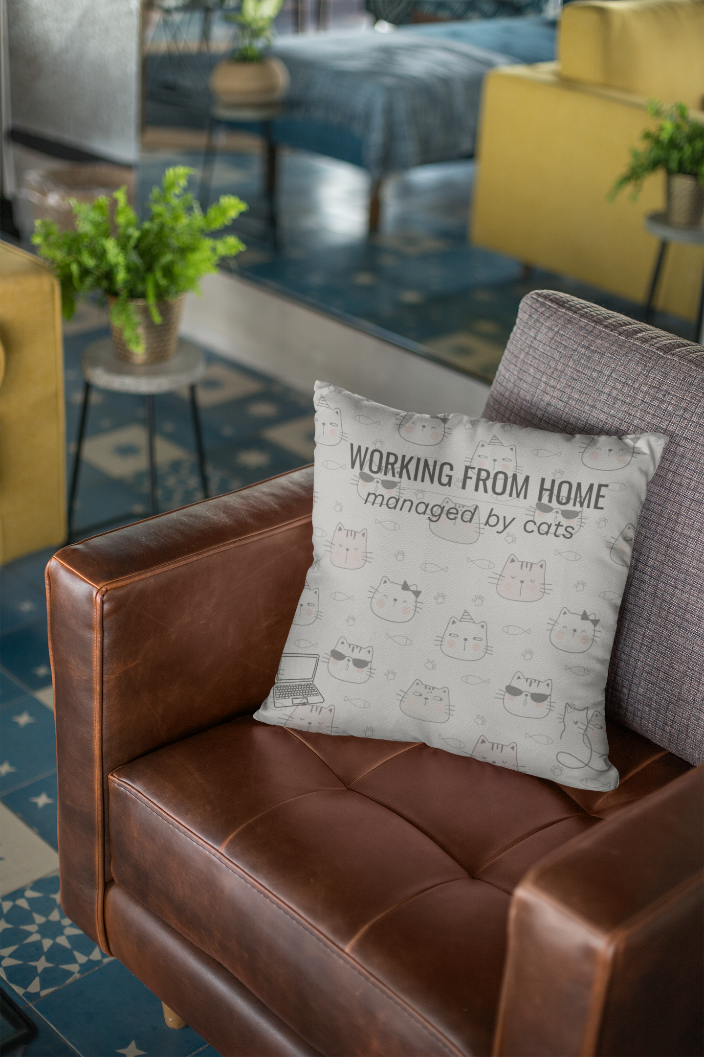 Working From Home Managed By Cats decorative throw pillow in light gray with cute cat doodle pattern, displayed on brown leather chair in modern home office setting with green plants in background