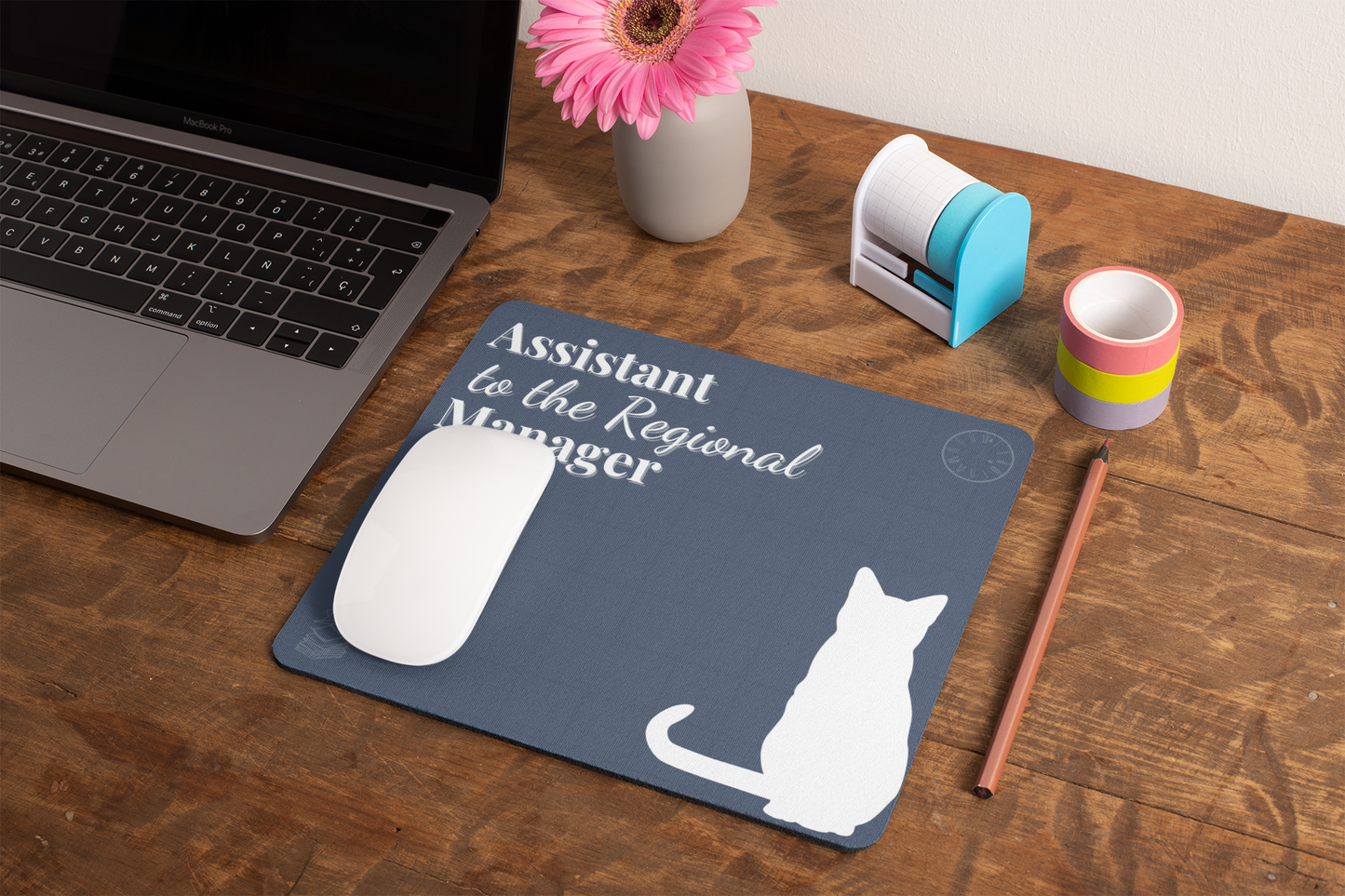 Assistant to the Regional Manager Cat Mousepad shown on wooden desk with laptop, pink gerbera daisy in vase, stamp holder, and colorful mug. Grey mousepad features white cat silhouette and office-themed text.