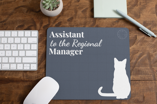 Assistant to the Regional Manager Cat Mousepad featuring a minimalist white cat silhouette on a slate grey background, displayed on a wooden desk with keyboard, mouse, succulent plant, and pen. Office-themed desk accessory with subtle clock detail.