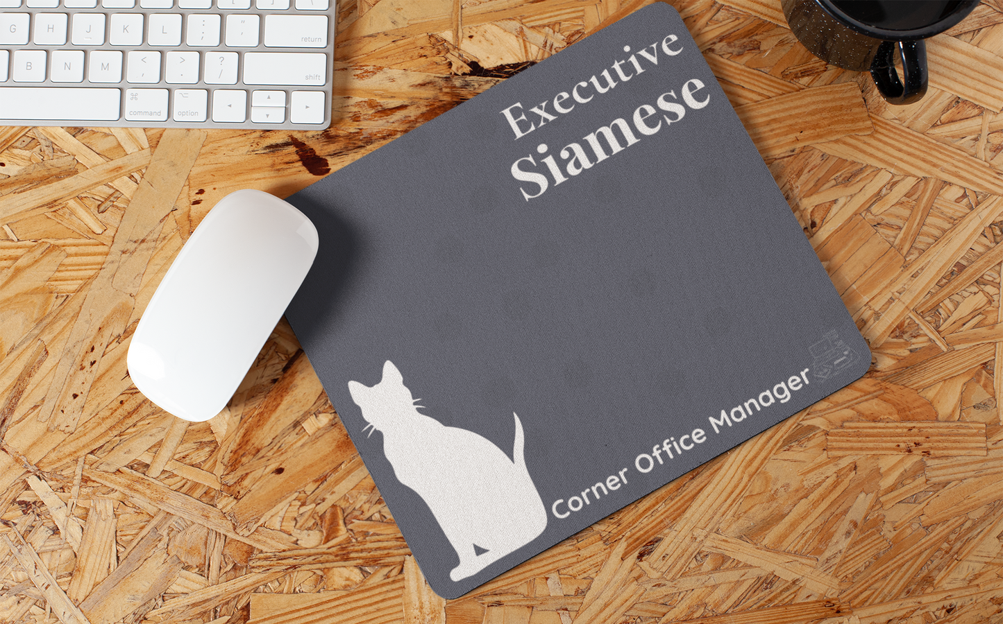 Executive Siamese - Corner Office Manager Mousepad featuring a slate blue background with white Siamese cat silhouette, displayed on wooden desk surface alongside white keyboard and black coffee mug. Non-slip design with elegant typography.