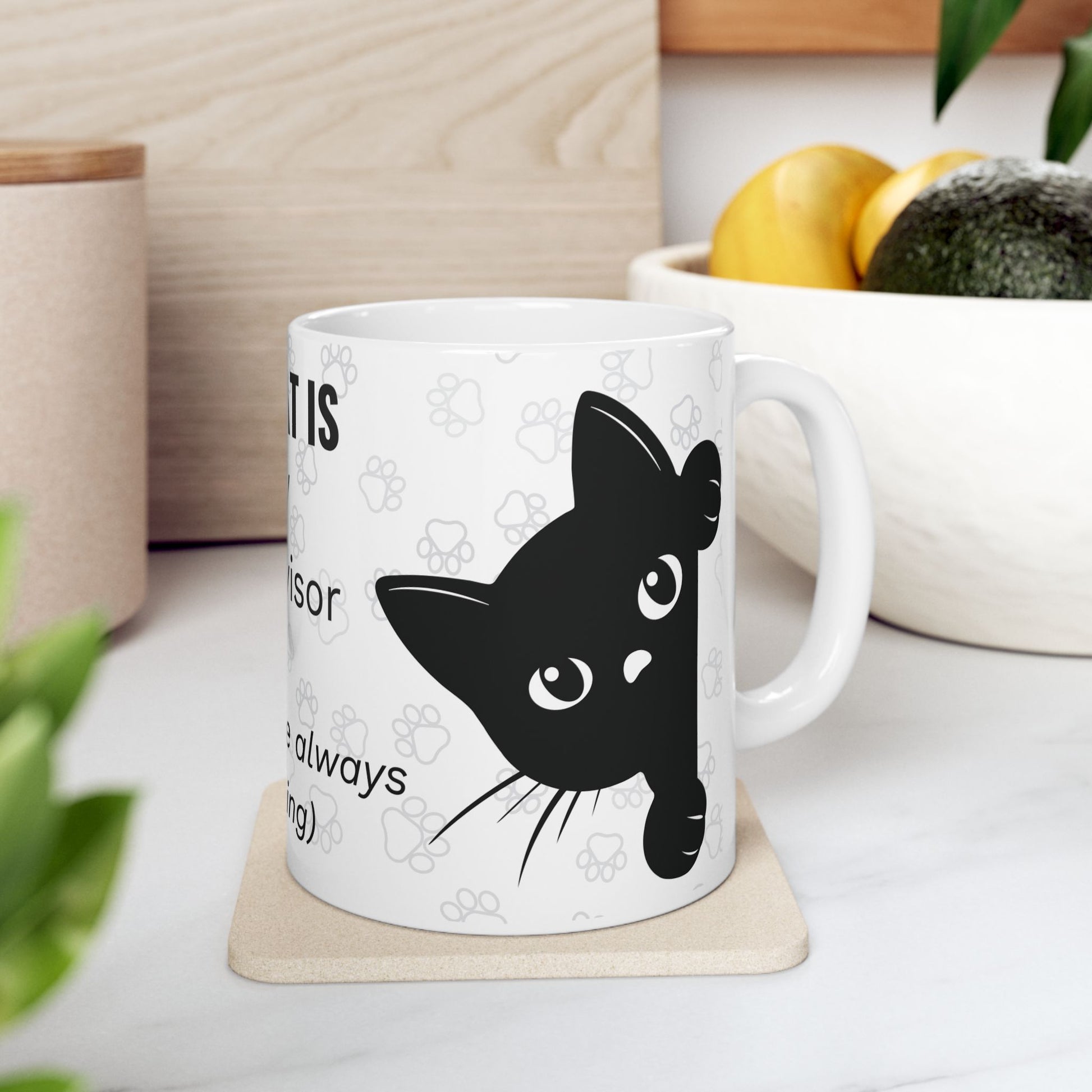 White ceramic mug featuring "THE CAT IS MY SUPERVISOR" text and black cat face design against paw print pattern background, showcasing a playful work-from-home theme with cat silhouette peeking from side, available in 11oz and 15oz sizes.