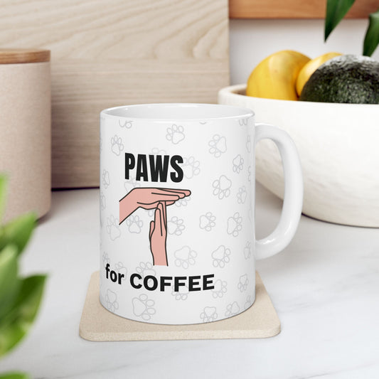 White ceramic mug displaying "Paws for Coffee" text with minimalist hand gesture and paw print pattern design, shown on beige coaster with fruit bowl and plants in background. Perfect Coffee Break Companion mug available in 11oz and 15oz sizes.